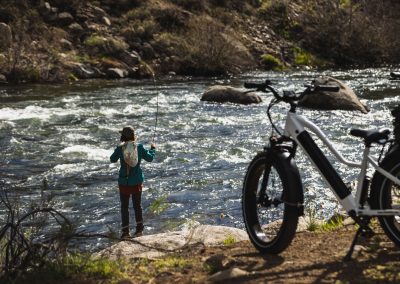 Woman standing on bank of river fly fishing with e-bike in foreground of shot.