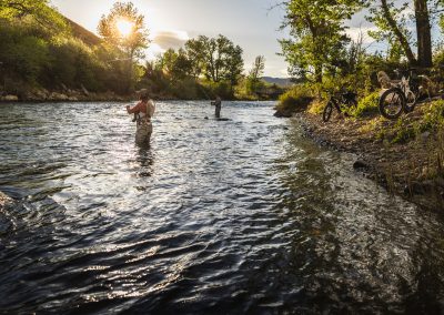 Scenic photograph of woman and man standing in the river fly fishing with e-bikes on shore.