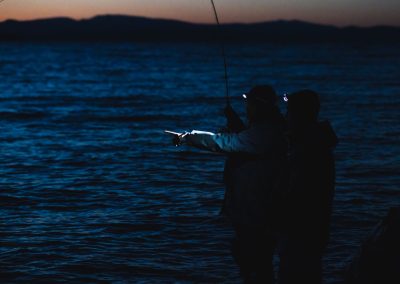 Photograph of two people fly fishing at dusk with headlamps.