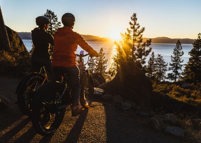 A couple riding e-bikes stoped on trail looking at view of lake and sunset.