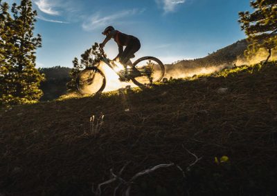 Female mountain biker riding dirt trail with sun setting behind her silhouette.